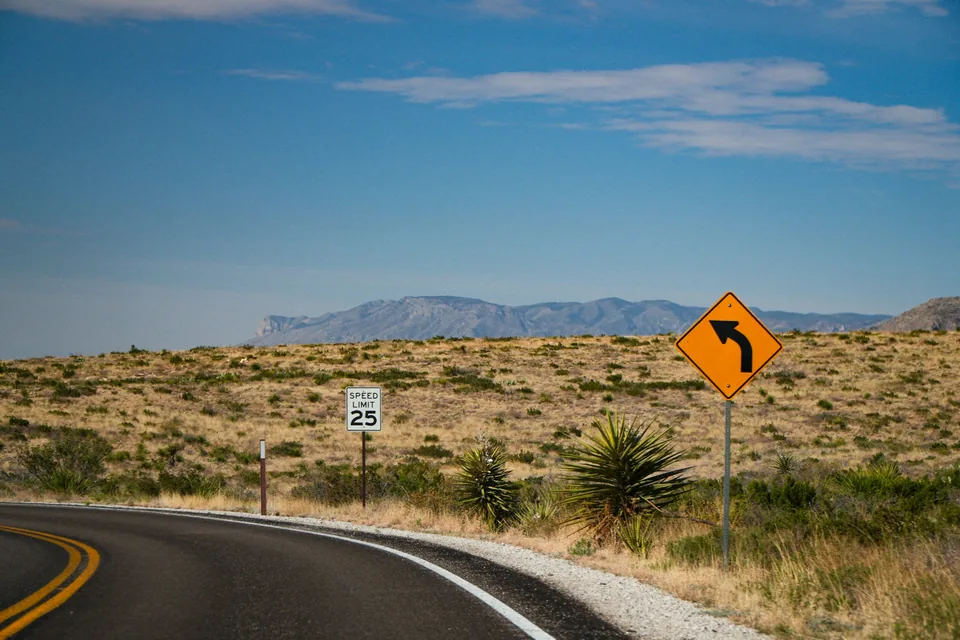 A two-lane road curving through dry grassland toward a distant mesa