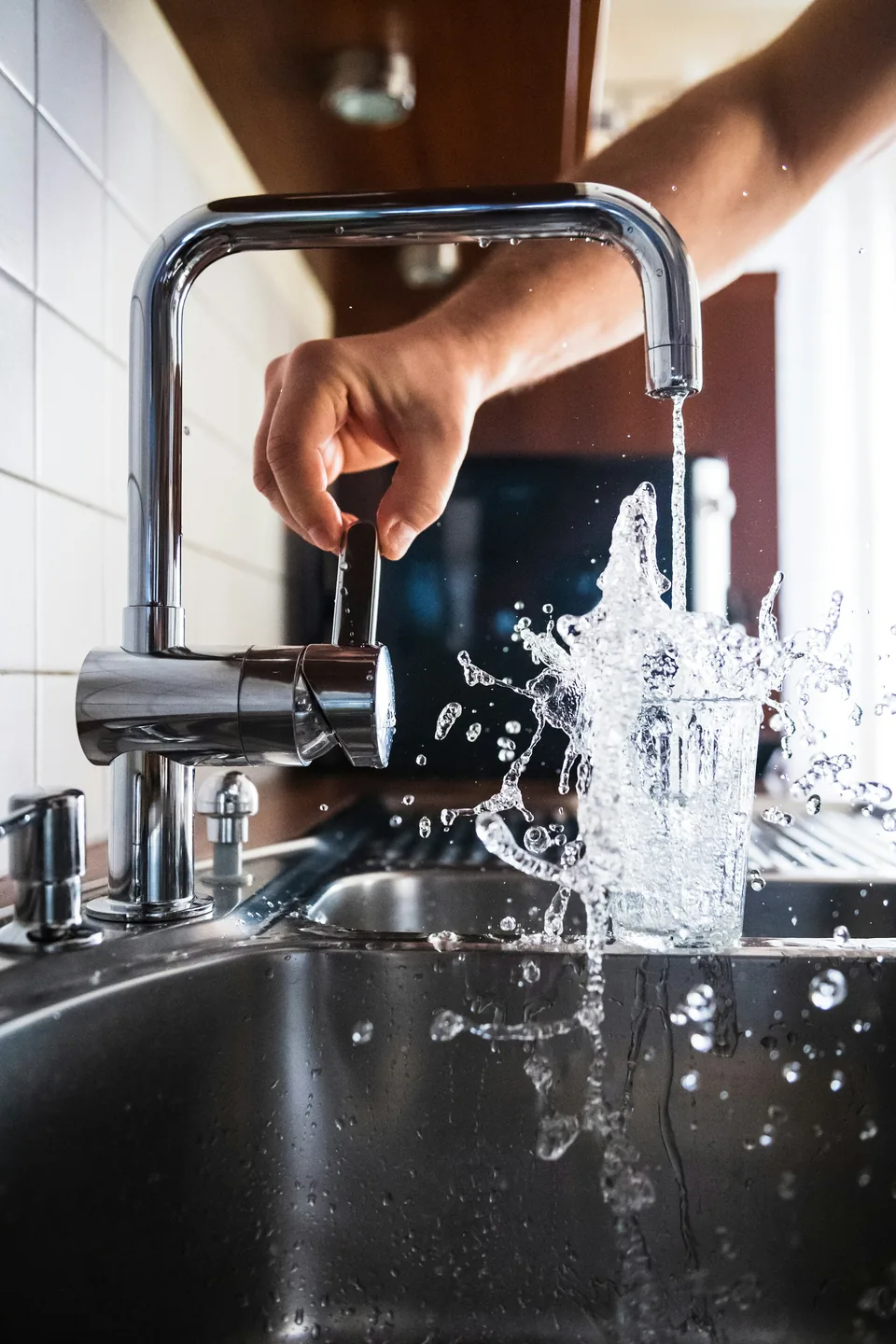 A hand turning a kitchen tap with water splashing into a steel sink