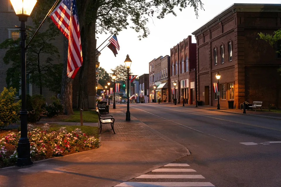 A small-town main street at dusk with a flag, street lamps and brick storefronts